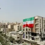 نمای دور از تهران که برج میلاد دیده می شود. در نمای نزدیکتر یک سمت ساختمان بلند تخریب شده و از سمت دیگر پرجم جمهوری اسلامی در ابعاد بزرگ نصب شده است