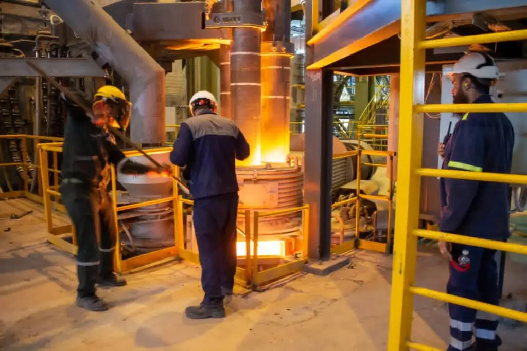 سه کارگر با لباس و کلاه ایمنی مشغول کار در کنار کوره ذوب فولاد. کوره کاملا داغ است و شعله های آتش و فولاد مذاب دیده می شود