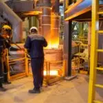 سه کارگر با لباس و کلاه ایمنی مشغول کار در کنار کوره ذوب فولاد. کوره کاملا داغ است و شعله های آتش و فولاد مذاب دیده می شود