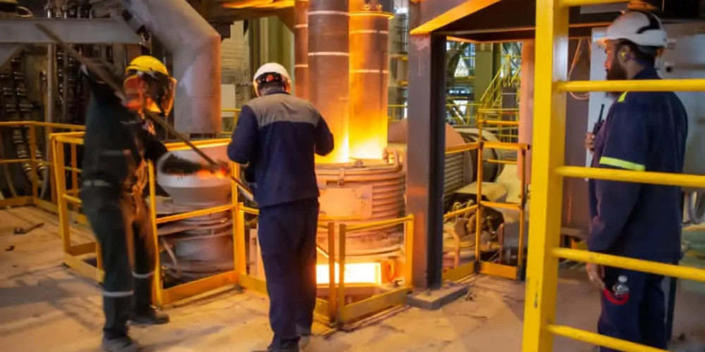 سه کارگر با لباس و کلاه ایمنی مشغول کار در کنار کوره ذوب فولاد. کوره کاملا داغ است و شعله های آتش و فولاد مذاب دیده می شود