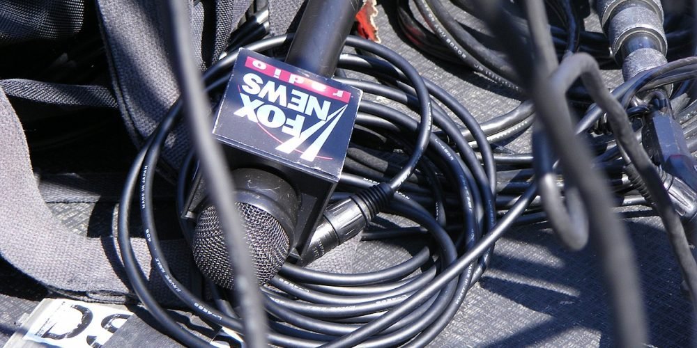 یک میکروفون مصاحبه که روی آن نوشته شده است فاکس نیوز. این میکروفون روی زمین است و سیم های سیاه رنگ زیادی اطراف آن ریخته شده است.