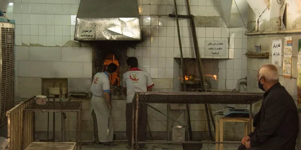 دو نانوا در حال پخت نان سنگک و یک پیرمرد مشتری دورتر از آنها نشسته است و منتظر است