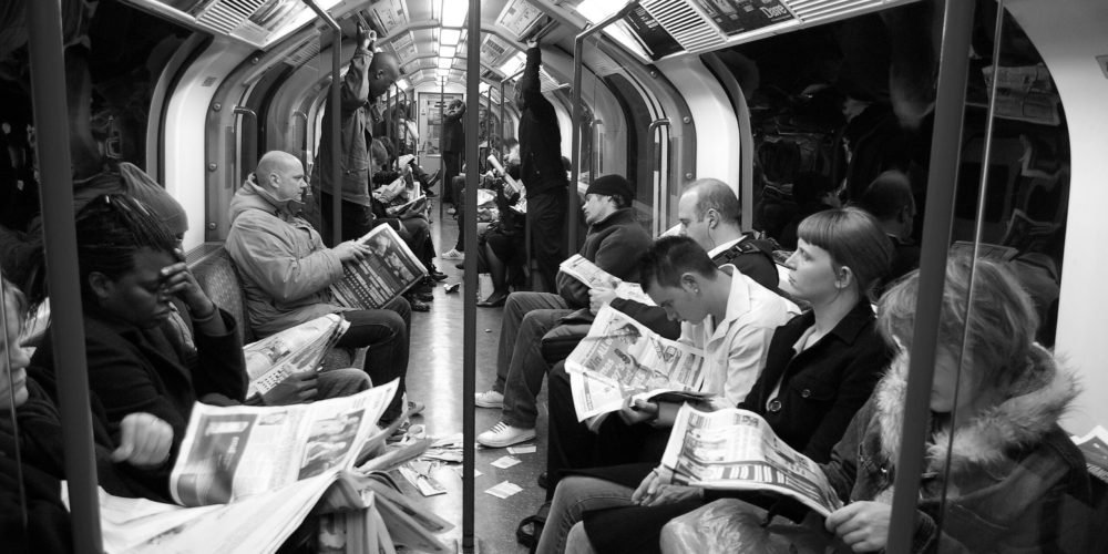 زن و مرد در دو سوی یک واگن مترو نشته اند و هر کس مشغول خواندن روزنامه است. عکس سیاه و سفید است